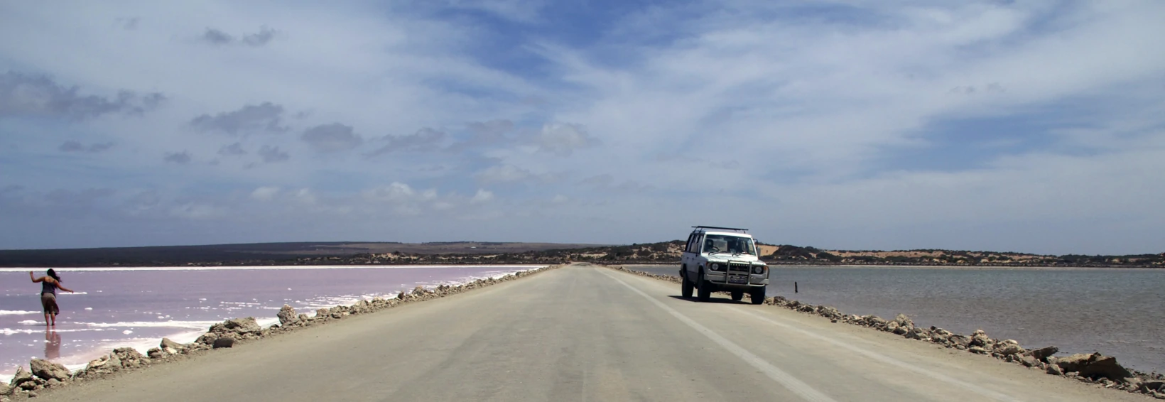 Dirt road with a single parked jeep on it. It divides two bodies of water, the one on the left is pink a woman is walking in it.